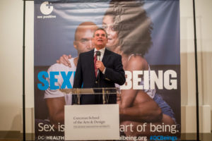 Man in suit with tie standing at podium speaking into microphone; large banner behind depicting couple sitting and "SEXUAL + BEING"