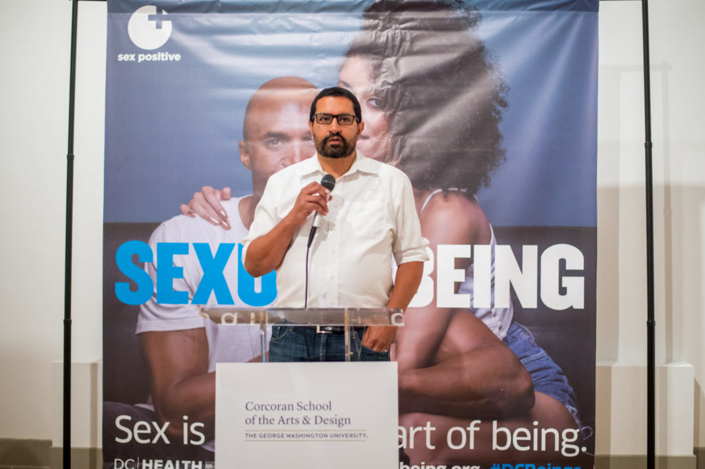 Man in white button-down shirt and jeans standing at podium, speaking into microphone; large banner behind depicting couple and "SEXUAL + BEING"