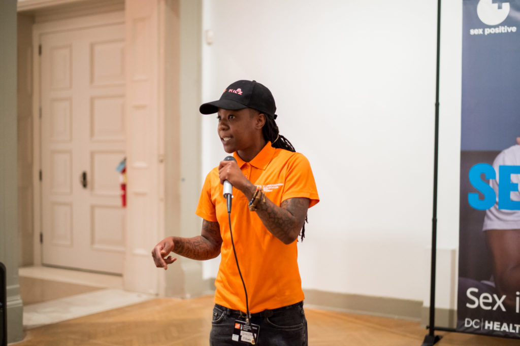 Woman wearing black hat and orange collared shirt speaks into microphone while gesturing with other hand.