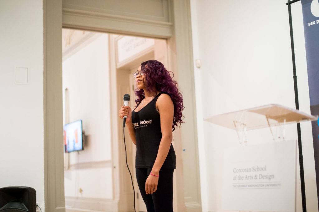 Woman with curly deep purple hair and glasses wearing black tank top hold microphone in front of podium.