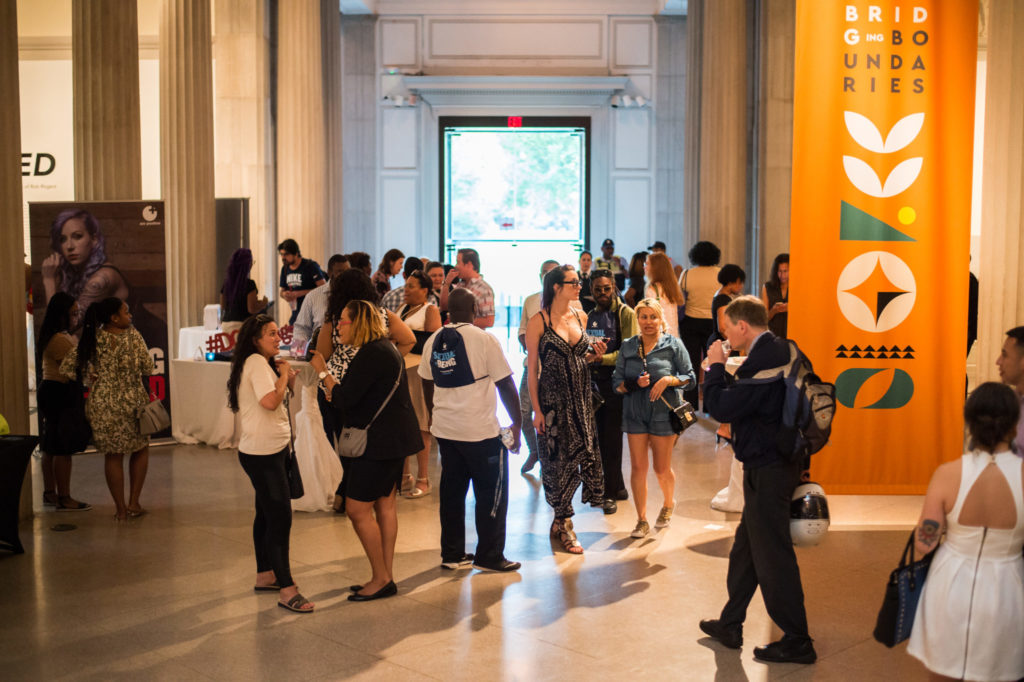 Diverse set of people in action, talking and walking through foyer with white columns and large banner depicting girl with purple hair.