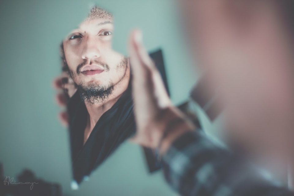 young black man with moustache and goatee holds broken shard of mirror and looks at self