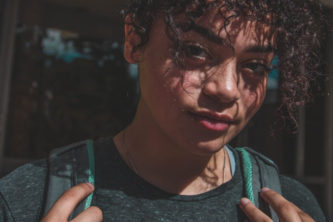 Young female with brown curly hair staring at camera, holding onto straps of gray and mint green backpack